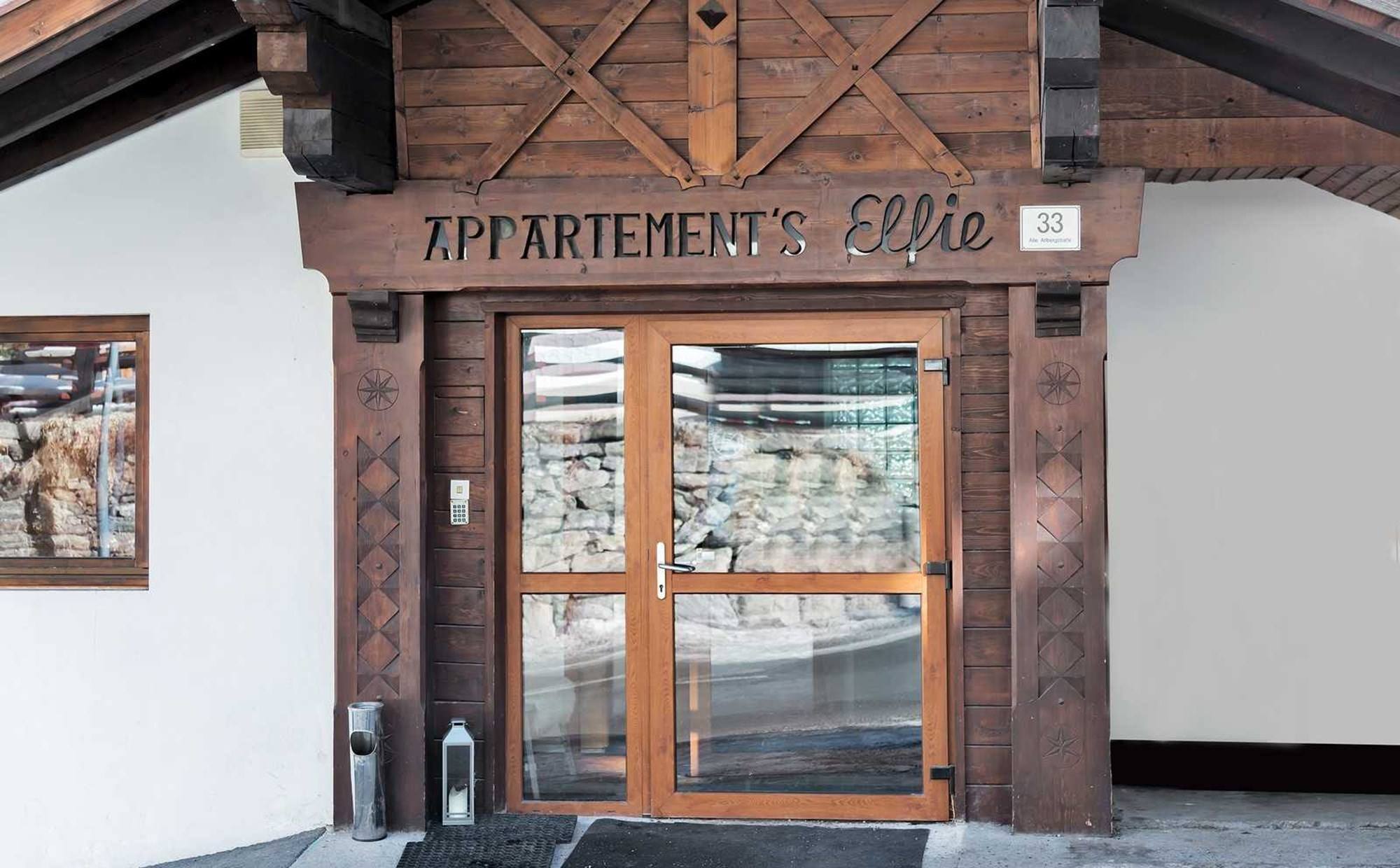 Bilden visar entrén till Appartements Ellie, ett av Thinggaards egna hotell. Det är en trädörr med glaspaneler, inramad i en rustik fasad. En skylt med namnet finns ovanför dörren med gatunumret 33 till höger. 