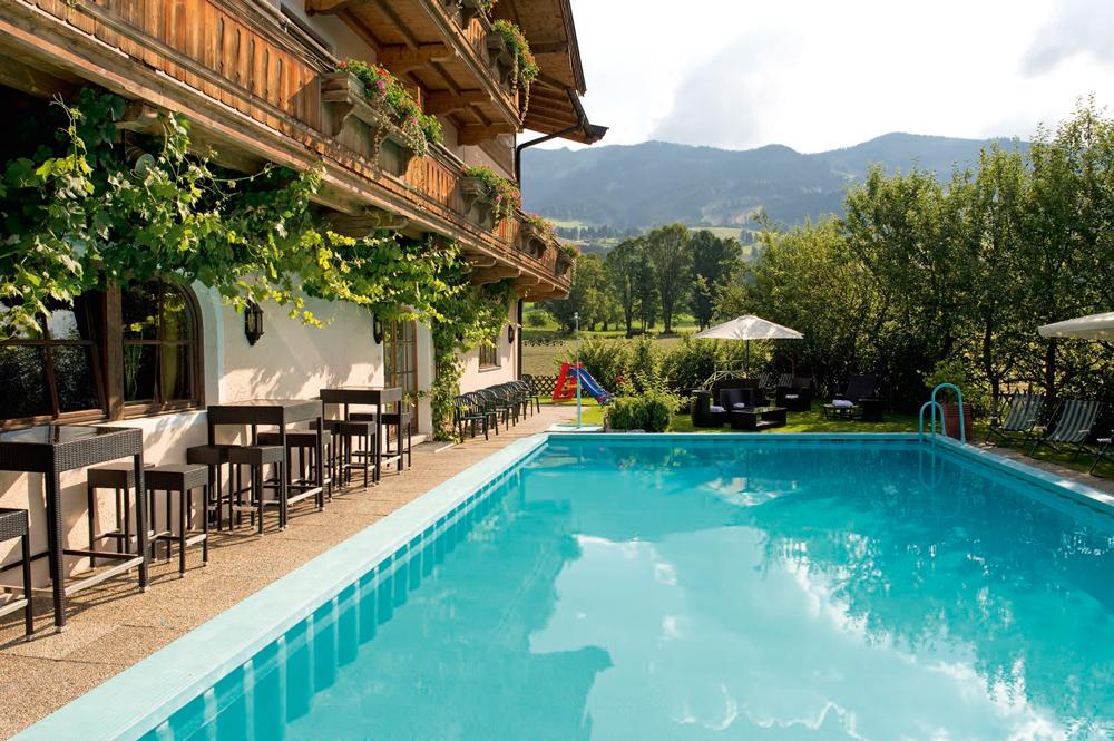 En rofylld utomhusmiljö i Westendorf med en klarblå pool kantad av solstolar och bord under parasoller. En rustik